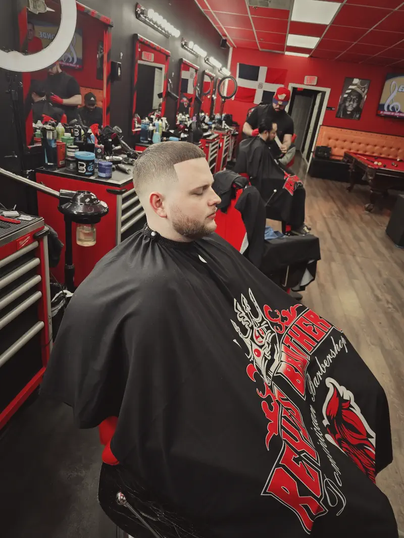 A client in a Reyes Brothers cape with a fresh skin fade, the row of stations and the Dominican flag on the back wall